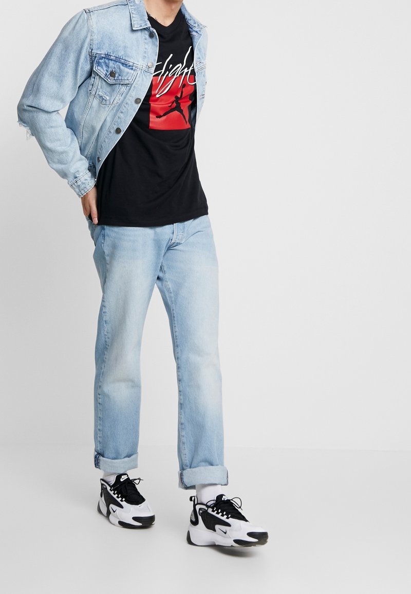 Persona que lleva puesta una chaqueta de mezclilla azul claro y jeans a juego, una camiseta negra con gráfico, calcetines blancos y zapatillas negras y blancas caminando.