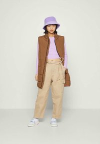 Colete acolchoado castanho, blusa de manga longa lavanda, calças cargo bege folgadas, chapéu roxo tipo balde e ténis brancos com toques de cores.