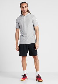Herre lysegrå polo-shirt med tre knapper, sorte shorts og farverige sportssko. Har logo og chevron-mønster på ærmerne.