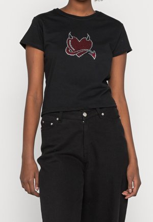 Osoba ubrana w czarny krótki top z czerwonym sercem i rogami diabła wykonanymi z kryształków, w zestawieniu z czarnymi, wysokim stanem spodniami.