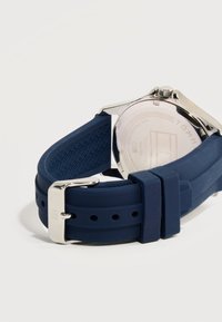 Πίσω μέρος ασημένιου ρολογιού με χαραγμένες λεπτομέρειες και λουράκι από υφασμένο σιλικόνη σε σκούρο μπλε χρώμα, στερεωμένο με ασημένια πόρπη.