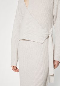 Robe pull en tricot côtelé beige clair avec un design enveloppant et une ceinture à la taille. Dotée d'un décolleté en V profond et de manches longues. Texture douce.