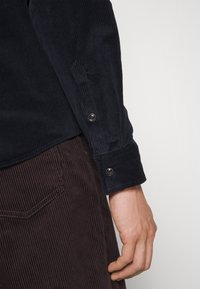 Granatowa koszula z sztruksu o fakturowanej powierzchni i mankietem na guziki, zestawiona z brązowymi spodniami ze sztruksu z pionowymi żebrowanymi wzorami.