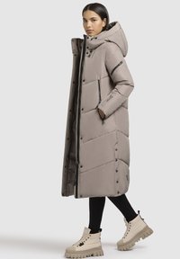 Gara, polsterēta mēteli gaisā bēžā krāsā, ar augstu apkakli, rāvējslēdzēju un pogu aizdari, priekšējiem kabatām un kapuci. Pārī ar apjomīgām zābaku podām.