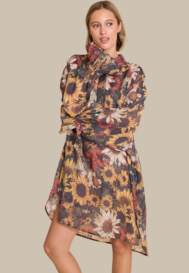 Blomstret kjole med lange ermer i et gjennomsiktig, lett stoff. Har en blanding av solfrø og andre blomster i varme toner på en mørk bakgrunn.