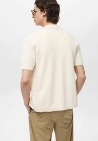 Maglione polo a maniche corte color crema, lavorato a maglia con texture, colletto piatto e orlo a coste, indossato con pantaloni beige chiaro. Vista posteriore.