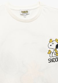 T-shirt en coton blanc avec un logo Snoopy noir et jaune sur le côté gauche de la poitrine. Encolure ronde avec une texture douce.