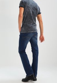 Jeans de mezclilla azul con corte recto, que presentan sutilezas en la costura y bolsillos traseros tradicionales. Llevados con una camisa corta de manga gris.