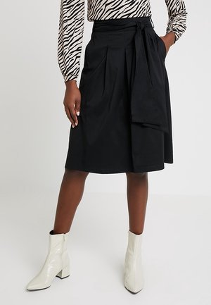 Sort midi-skjørt med plisseringer og et bundet stofbælte i taljen. Materialet ser glat og let ud. Båret med hvide ankelstøvler.