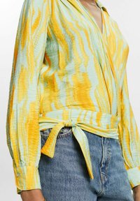 Blouse à manches longues avec un motif abstrait bleu et jaune. Fabriquée en tissu léger avec une taille nouée et des poignets boutonnés.