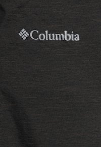 Columbia logo tumšā pelēkā teksturētā audumā, kurā ir smalks raksts ar brūnajiem plankumiem. Izteikta zīmola emblēma, iegriezta audumā.