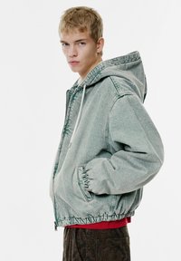 Jachetă din denim de culoare bleu deschis, cu glugă. Dispune de manșete și tiv elastice, închidere cu fermoar și șnur de ajustare. Pur și simplu peste o cămașă roșie și pantaloni cu model.