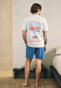 Mand stående barfodet i blå shorts og en hvid T-shirt med en kaffemaskinegrafik og tekst på bagsiden i et soveværelsesmiljø.