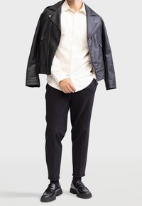 Veste de motard en cuir noir portée sur une chemise blanche à boutons, associée à un pantalon noir fuselé et des mocassins noirs. Texture lisse, coupe ajustée.