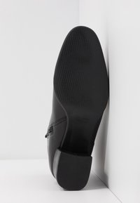 Chaussure en cuir noir à surface lisse, avec un talon façonné et une semelle en caoutchouc texturée, affichant l'étiquette de taille "38" sous la semelle.