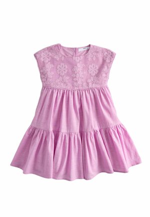 Joven niña de pie con un vestido rosa bordado con flores y sandalias beige con tiras texturizadas sobre un fondo liso.