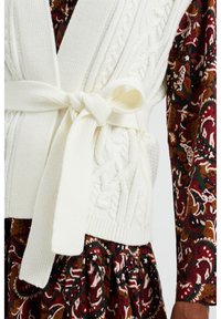 Gilet en tricot blanc avec des motifs en relief et une ceinture nouée, associé à une robe à motifs floraux bordeaux et verts. Tissu texturé et doux.