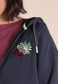 Sweat à capuche bleu marine présentant un motif brodé vert et rouge sur la poitrine, avec des accents verts contrastants sur la capuche et la fermeture éclair.
