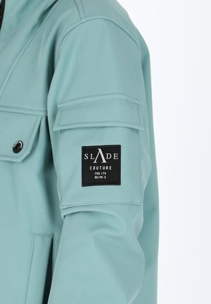 Aquamarinfarbige Jacke aus glattem Stoff, ausgestattet mit einem Patch mit "SLADE COUTURE" in Weiß auf schwarzem Hintergrund und einer Fronttasche.