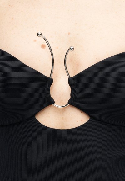 Tissu noir côtelé relié par un fermoir métallique incurvé argenté avec deux extrémités en forme de boule, montrant une peau pâle avec un grain de beauté en arrière-plan.