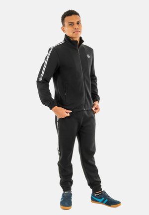 Chándal negro de tejido suave, con rayas blancas a lo largo de las mangas y los laterales. Chaqueta con cremallera, puños elásticos y dos bolsillos laterales.