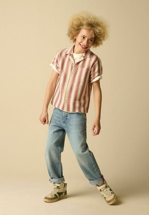 Kind met krullend haar dat een kort mouwenshirt met patroon, opgerolde spijkerbroek en sneakers draagt, staat glimlachend voor een beige achtergrond.