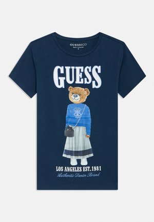 Marineblå t-skjorte med stor "GUESS"-tekst, teddybjørn som har på seg en blå genser, grått skjørt, hvite sko og holder en liten svart veske.