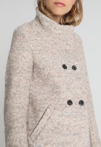 Cappotto in misto lana beige con finitura testurizzata, colletto alto, bottoni neri e tasche laterali. Design doppiopetto.