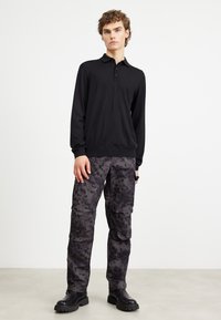 Camisa pólo preta de manga longa com três botões, combinada com calças cargo camufladas escuras, com bolsos e um acabamento texturizado, usadas com botas pretas.