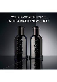 Deux flacons de parfum noirs Hugo Boss avec logos embossés sont posés sur une surface réfléchissante sous le texte à propos d'un tout nouveau logo.
