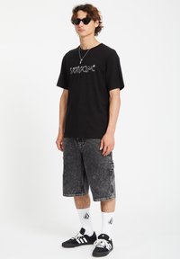 Black t-shirt with white "Volcom" logo, gray denim cargo shorts, black sneakers with white accents, and white socks with a logo.