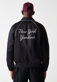 Juoda satino striukė su guminiu rankogaliu ir apatine dalimi, balta "New York Yankees" siuvinėjimu ant nugaros, suapvalintomis pečiomis ir subtiliu dizainu.