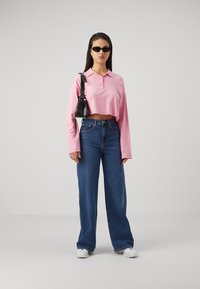 Pink, cropped long-sleeve polo shirt with a collar and button detail, paired with blue wide-leg jeans and white sneakers. Black bag and sunglasses.