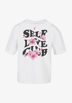 Bílá košile s krátkým rukávem se silným černým nápisem "SELF LOVE CLUB" a růžovými květy sakura rozptýlenými kolem nápisu.