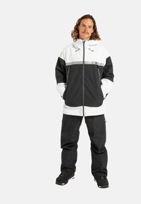 Zwart-witte waterdichte jas met verstelbare capuchon, grijze streep en ritszakken, gecombineerd met bijpassende zwarte broek en zwart schoeisel.