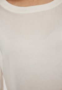 Fehér kötött pulóver, széles, kerek nyakkivágással és finom bordázott textúrával. Az anyag puha és enyhén áttetsző.