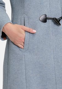 Lichtblauwe wollen jas met een gestructureerde stof, schuine zakken en zwarte sluiting met toggle-detail. Hand gedeeltelijk zichtbaar in de zak.