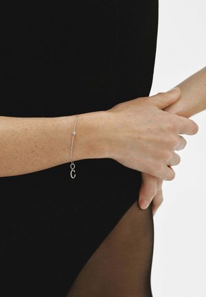 Persona sosteniendo una muñeca con la otra mano, llevando una delicada pulsera de plata con un pequeño charm, vestida con ropa negra.