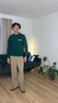 Mladi moški s kodrastimi lasmi, ki nosi zelen pulover in bež hlače, stoji na lesenem tleh v sodobni dnevni sobi z rastlinami in kavčem.