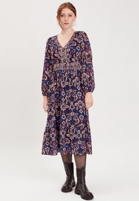 Largo vestido morado con estampado floral en azul y crema. Escote en V, mangas largas abullonadas y cintura fruncida. Llevado con botines negros hasta el tobillo.