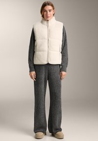 Hellbeige gesteppte Fleeceweste mit hohem Kragen, Frontreißverschluss und Seitentaschen. Kombiniert mit grauen, weit geschnittenen Strickhosen und beigen Slip-On-Schuhen.