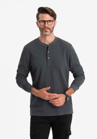 Mann mit Brille, der ein dunkelgraues Langarm-Henley-Shirt und schwarze Hosen trägt, steht mit locker gefalteten Händen.