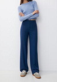 Une femme se tient debout avec les bras croisés, portant un haut à manches longues à rayures bleu clair, un pantalon large bleu foncé et des baskets grises, sur fond blanc.