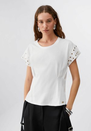 Donna con capelli lunghi castani che indossa una maglietta bianca a maniche corte con occhielli metallici sulle maniche e pantaloni neri, accessoriata con orecchini e bracciale in argento.