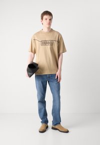 Beige bomull t-skjorte med svart "HUGO" logo, paret med blå denimjeans. Holder en svart tilbehør. Lysbrune semskede sko, flat såle.