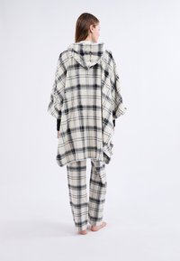 Poncho à carreaux en crème et noir, avec capuche et fentes latérales, associé à un pantalon à carreaux assorti. Tissu doux et douillet avec une coupe décontractée.