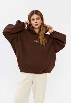 Bruine oversized hoodie met een trekkoordcapuchon, geribbelde manchetten en een cinched zoom. Bevat een klein gouden logo op de borst.
