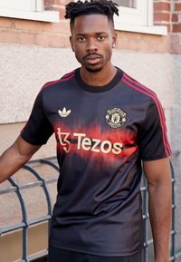 Camiseta deportiva negra con acentos rojos, con un diseño degradado, logo de "Tezos" en dorado y el escudo del Manchester United en el pecho.