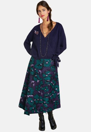 Marineblauer Strickpullover mit V-Ausschnitt und seitlichem Bindeband, kombiniert mit einem langen lila Rock mit grünen Blumenmustern und schwarzen Schnürstiefeln.