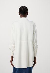 Camicia bianca a maniche lunghe con colletto strutturato, caratterizzata da una cucitura posteriore, polsini con bottoni e una vestibilità ampia, realizzata in un tessuto morbido.